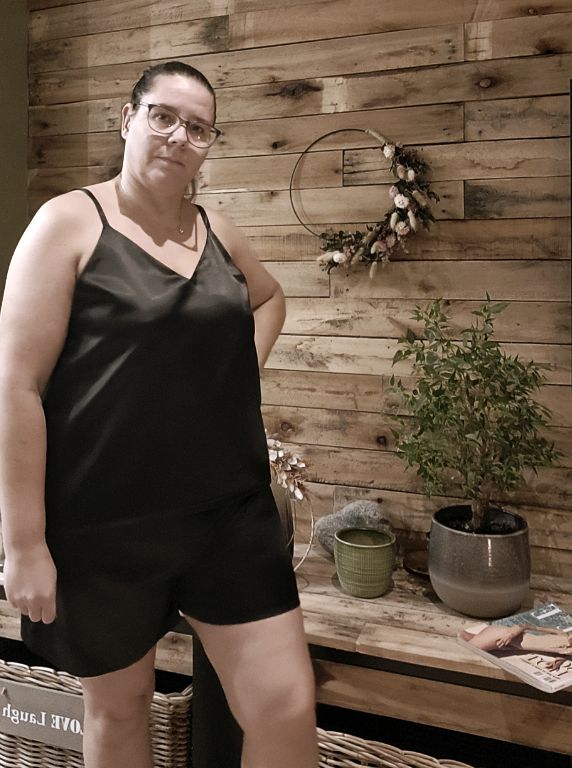 Close-up van een zijdezachte satijnen Aurora pyjama in zacht glanzend materiaal, gedragen door een vrouw die ontspannen zit, met subtiele details en een elegante pasvorm. Aurora pyjama  met spaghettitop, lange broek of short.

